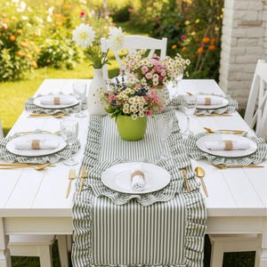 Peut inclure: Une table en bois blanche dress&eacute;e pour un repas en plein air. La table est orn&eacute;e d'un chemin de table et de sets de table &agrave; rayures vertes et blanches. Des assiettes blanches, des couverts dor&eacute;s et des verres sont dispos&eacute;s.