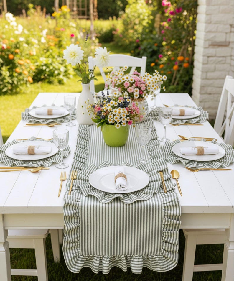 Peut inclure: Une table en bois blanche dress&eacute;e pour un repas en plein air. La table est orn&eacute;e d'un chemin de table et de sets de table &agrave; rayures vertes et blanches. Des assiettes blanches, des couverts dor&eacute;s et des verres sont dispos&eacute;s.