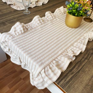 May include: A beige and white striped table runner with ruffled edges. The runner is rectangular and features vertical stripes. A gold-colored pot with a plant is on the right side of the runner.