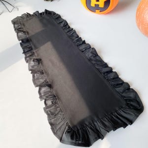 May include: A long, rectangular black table runner with ruffled edges. The runner is made of a smooth, dark fabric and is decorated with a delicate, ruffled trim. The table runner is suitable for Halloween or other special occasions.