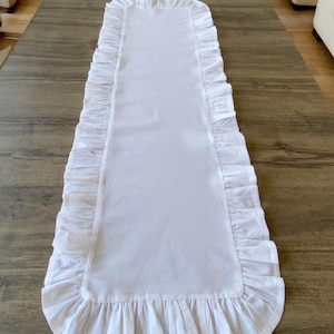 May include: A white rectangular table runner with ruffled edges. The runner is centered on a dark wooden table. The ruffles are along the long sides and the ends of the runner. The runner is made of a soft, white fabric.