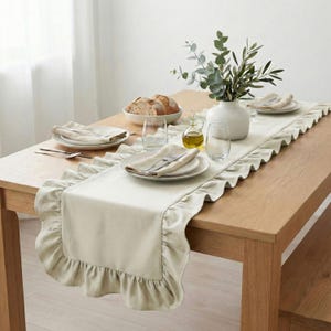 May include: A beige linen table runner with a ruffled edge is displayed on a wooden table. The table is set with white plates, silverware, napkins, and glasses. A white vase with greenery and a bowl of bread complete the dining scene.