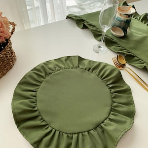 May include: An olive green placemat with a ruffled edge, set on a white table. A matching runner with ruffled edges is also visible. Gold-coloured cutlery, a wine glass, and a decorative cup are also on the table. A wicker basket with pink flowers is in the background.
