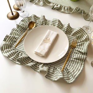 May include: A place setting featuring a white plate, a folded white napkin with gold embroidery, and gold cutlery. The plate rests on a green and white striped placemat with a ruffled edge. A matching table runner is visible in the background.