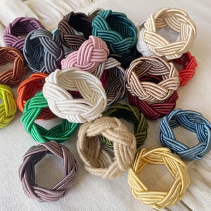 May include: A collection of braided fabric napkin rings in various colors, including white, green, red, and brown. The rings are arranged on a white surface, showcasing their woven texture and diverse hues.