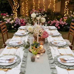 Peut inclure: Une table de salle &agrave; manger ext&eacute;rieure joliment dress&eacute;e avec des assiettes blanches, des couverts dor&eacute;s et des verres &agrave; vin. La table est d&eacute;cor&eacute;e d'un chemin de table &agrave; rayures vertes et blanches, de centres de table floraux et de bougies. Des guirlandes lumineuses pendent en arri&egrave;re-plan.