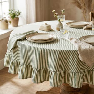 May include: A round table set for dining, featuring a green and white striped tablecloth with a ruffled edge. The table is set with plates, napkins, glasses, and a butter dish. The table is made of wood.