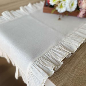 May include: Cream-coloured rectangular table runner with frilled edges. The runner is made of a textured fabric and features delicate stitching along the frills. A wooden tray with flowers sits in the background.