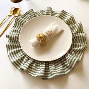 May include: A white plate with a folded white napkin and gold napkin ring sits atop a green and white striped placemat. Gold utensils are to the left. The napkin has gold embroidered script. The placemat has a ruffled edge.