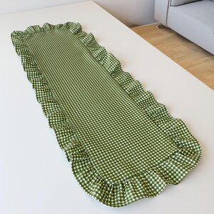 May include: A green and white gingham table runner with frilled edges. The runner has a rectangular shape and a classic checkered pattern. The frilled edges provide a decorative element to the table setting.