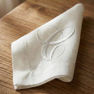 May include: A folded white linen napkin featuring an embroidered capital letter "C" in a script style. The napkin has a hemmed edge and is placed on a wooden surface. The embroidery thread matches the napkin's color.