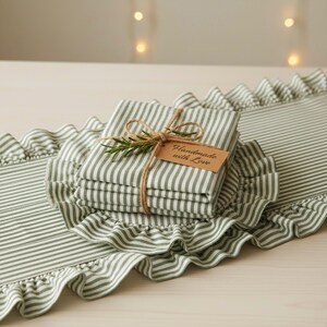 Peut inclure: Un ensemble de serviettes pli&eacute;es &agrave; rayures vertes et blanches, attach&eacute;es avec de la ficelle et une branche de romarin. Une &eacute;tiquette indique "Handmade with Love". Les serviettes sont pos&eacute;es sur un chemin de table assorti &agrave; volants.