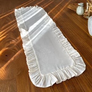 May include: A white table runner with a ruffled border. The runner is made of a soft, linen-like fabric and is perfect for adding a touch of elegance to any table setting.