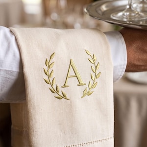 May include: A cream-colored linen napkin featuring a gold embroidered laurel wreath design surrounding the letter A in the center. The fabric has a visible woven texture and a decorative hemstitch detail along the bottom edge of the cloth.