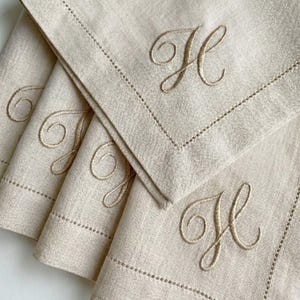 May include: A stack of four beige linen napkins. Each napkin features an embroidered capital letter "H" in a matching beige thread. The edges are finished with a decorative stitched border, adding a touch of elegance to the simple design.