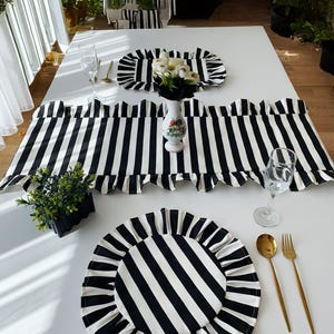 May include: A table setting with black and white striped placemats, a table runner, and matching plates. Gold-colored cutlery and clear wine glasses are arranged on a white table. A small vase with white flowers is in the center.