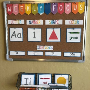 May include: A corkboard with the words "WEEKLY FOCUS" in colourful letters at the top. Below are four categories: "LETTER", "NUMBER", "SHAPE", and "COLOR". Each category has a corresponding image: the letter "Aa", the number "1", a red triangle, and a green crayon. Below the categories is a row of four sight words: "a", "at", "in", and "can".