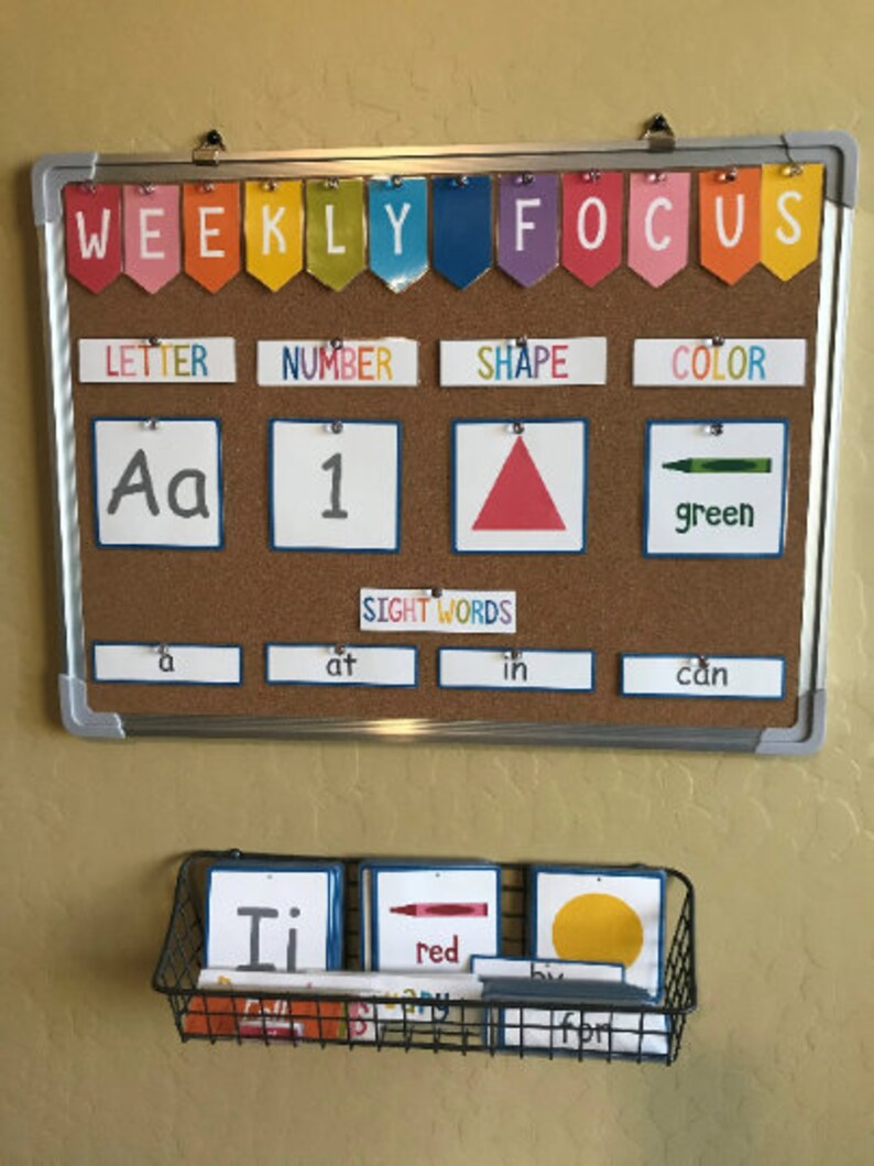 May include: A corkboard with the words "WEEKLY FOCUS" in colourful letters at the top. Below are four categories: "LETTER", "NUMBER", "SHAPE", and "COLOR". Each category has a corresponding image: the letter "Aa", the number "1", a red triangle, and a green crayon. Below the categories is a row of four sight words: "a", "at", "in", and "can".