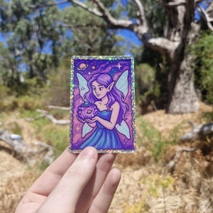 May include: A holographic sticker featuring a purple-haired fairy with light blue wings, holding a cosmic orb. The background is a starry purple sky with a planet and stars. The fairy wears a blue dress.
