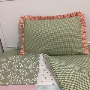 May include: A green and white gingham pillow with an orange frilled edge sits on a patchwork quilt. The quilt features a variety of patterns, including a floral design, small dots, and more gingham.