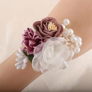 May include: A wrist corsage with mauve and white fabric flowers, and faux pearls. The corsage has small white floral accents and green leaves. The flowers are attached to a wrist band.
