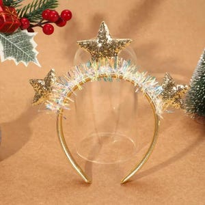 Puede incluir: Diadema dorada con tres estrellas brillantes. Las estrellas son doradas y la diadema tiene un adorno similar a oropel. Un accesorio festivo, ideal para celebraciones navideñas o eventos temáticos.