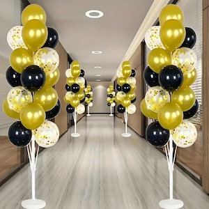 Pode incluir: Um corredor decorado com buquês de balões. Cada buquê apresenta balões dourados, pretos e cheios de confetes. Os balões são dispostos em suportes brancos, criando um ambiente festivo e comemorativo.