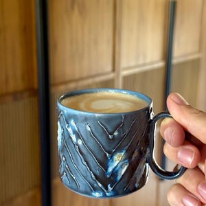 Puede incluir: Una taza de cerámica azul con un patrón de chevrón texturizado, llena de una bebida cremosa. La taza tiene un asa y un borde azul oscuro, y el interior está lleno de un líquido marrón claro. La taza se sostiene en una mano.