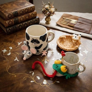May include: A ceramic mug with a black and white cow pattern featuring a crocheted grey mouse attachment, a smaller ceramic mug with a crocheted green frog attachment, a round crocheted tan coaster, and a set of metal crochet hooks in a leather case.