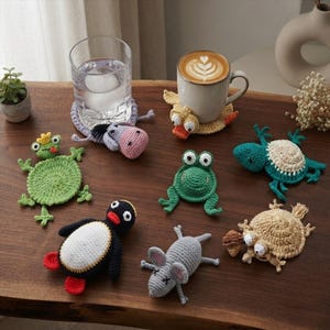 Può includere: Una collezione di sette sottobicchieri all'uncinetto fatti a mano a forma di vari animali, tra cui una rana verde con corona, un asino grigio, un'anatra gialla, una rana verde, una tartaruga verde acqua, un pinguino bianco e nero e un topo grigio.