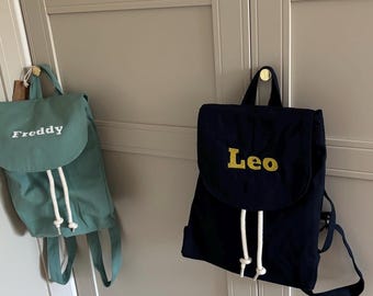 May include: Two personalised backpacks. One is teal with "Freddy" embroidered in white, the other is navy blue with "Leo" embroidered in gold. Both have drawstring closures and adjustable straps.
