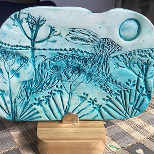 Könnte beinhalten: Hellblaue Keramikplatte mit einem Hasen, der durch ein Feld rennt, mit einem Vollmond. Das Kunstwerk ist auf einem Holzständer montiert. Die Platte hat dunkelblaue Details und zeigt eine Naturszene mit Bäumen und Pflanzen.