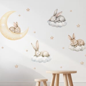 Op de afbeelding: Een set muurstickers met aquarelillustraties van lichtbruine konijnen die slapen op een gele maansikkel en pluizige witte wolken, geaccentueerd door kleine beige sterren die verspreid zijn voor een hemels tafereel.