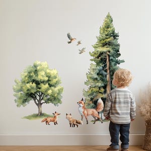 Könnte beinhalten: Ein Wandaufkleber-Set mit einer Waldszene im Aquarellstil, bestehend aus einem großen grünen Laubbaum, einer hohen immergrünen Tanne, drei kleinen Füchsen in Orange und Weiß sowie drei kleinen Vögeln, die über den Tieren in der Luft fliegen.
