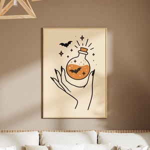May include: A framed print featuring a hand holding a potion bottle with a bat and stars. The artwork is in a minimalist style with black line art on a beige background. The print is displayed in a living room setting with a sofa, coffee table, and decorative plants.