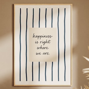 May include: A framed print with a cream background and navy blue vertical stripes. A smaller cream-coloured rectangle in the centre features the text "happiness is right where we are" in a handwritten font. Dried plants are in the lower right corner.
