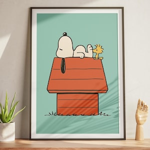 May include: Framed art print depicting Snoopy and Woodstock on Snoopy's doghouse. The illustration has a light blue background with a red doghouse and green grass. Snoopy is white, and Woodstock is yellow. The artwork is displayed on a wooden cabinet.