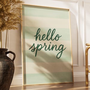 May include: A framed art print with the words "hello spring" in green script on a striped background of light green and beige. A large, dark brown vase with dried plants is to the left, and a small bamboo table is to the right.