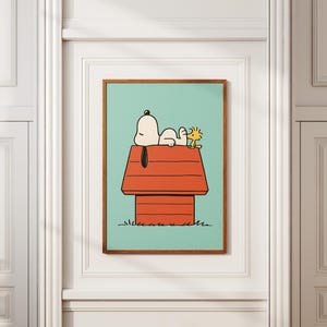 May include: A framed art print depicting Snoopy and Woodstock. Snoopy is resting on his red doghouse, with Woodstock beside him. The background is a light teal, and the frame is brown.