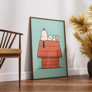 May include: Framed art print depicting Snoopy and Woodstock. Snoopy is lying on his red doghouse, with Woodstock beside him, set against a light blue backdrop. The artwork is in a wooden frame and leans against a white wall.