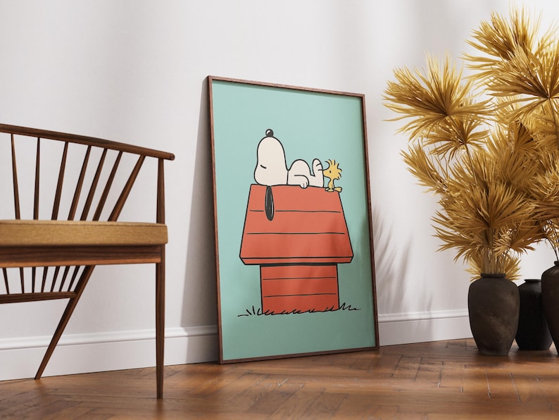 May include: Framed art print depicting Snoopy and Woodstock. Snoopy is lying on his red doghouse, with Woodstock beside him, set against a light blue backdrop. The artwork is in a wooden frame and leans against a white wall.