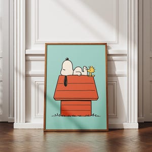 May include: A framed art print depicting Snoopy and Woodstock on Snoopy's red doghouse against a light blue backdrop. The artwork is in a cartoon style, with a brown wooden frame. The background is a white wall.