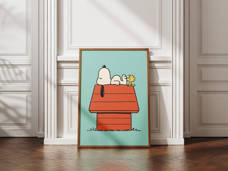 May include: A framed art print depicting Snoopy and Woodstock on Snoopy's red doghouse against a light blue backdrop. The artwork is in a cartoon style, with a brown wooden frame. The background is a white wall.