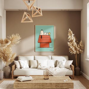 May include: A living room with a framed Snoopy art print above a cream-colored sofa with cushions. The room has a neutral colour scheme with wooden features, including a coffee table and geometric light fittings. Tall, dried plants in vases add texture.