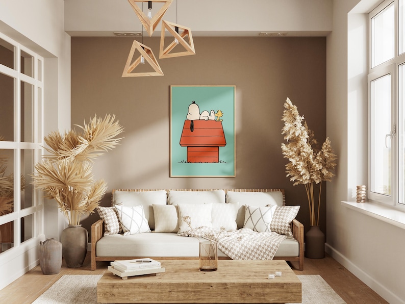 May include: A living room with a framed Snoopy art print above a cream-colored sofa with cushions. The room has a neutral colour scheme with wooden features, including a coffee table and geometric light fittings. Tall, dried plants in vases add texture.