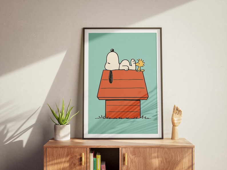 May include: Framed art print depicting Snoopy and Woodstock on Snoopy's doghouse. The illustration has a light blue background with a red doghouse and green grass. Snoopy is white, and Woodstock is yellow. The artwork is displayed on a wooden cabinet.