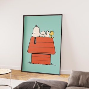 May include: A framed print featuring Snoopy and Woodstock. Snoopy is lying on his red doghouse, with Woodstock beside him. The background is a light teal colour, and the frame is black.