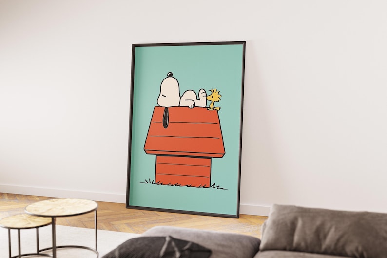 May include: A framed print featuring Snoopy and Woodstock. Snoopy is lying on his red doghouse, with Woodstock beside him. The background is a light teal colour, and the frame is black.