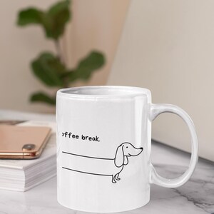 Könnte beinhalten: Weiße Keramik-Kaffeetasse mit einer schwarzen Strichzeichnung eines Dackelhundes und dem Schriftzug "coffee break". Die Tasse hat einen gebogenen Henkel und steht auf einer weißen Oberfläche.