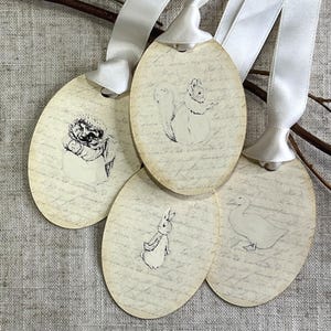 Könnte beinhalten: Vier ovale Geschenkanhänger mit weißen Satinbändern. Jeder Anhänger zeigt eine andere Tierzeichnung: ein Igel, ein Eichhörnchen, ein Kaninchen und eine Ente. Die Anhänger haben ein Vintage-Aussehen aus gealtertem Papier mit Schrift.